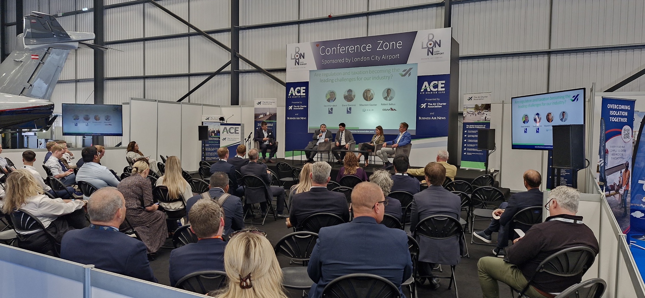 Delegates watching a panel discussion with a branded stage, projection and audience screens at Air Charter Expo