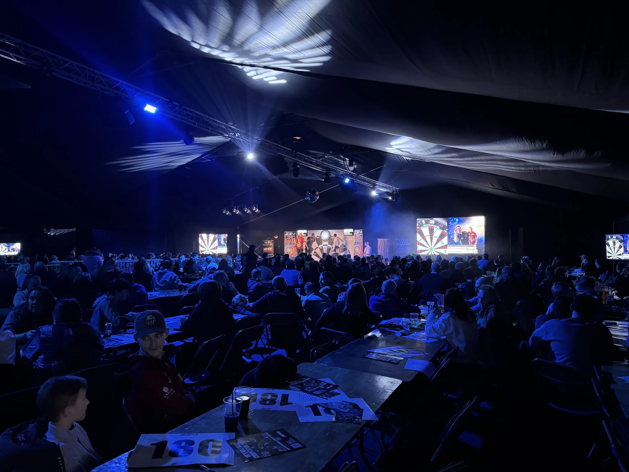 Audience watching a live darts event with blue show lighting, a stage and large screens