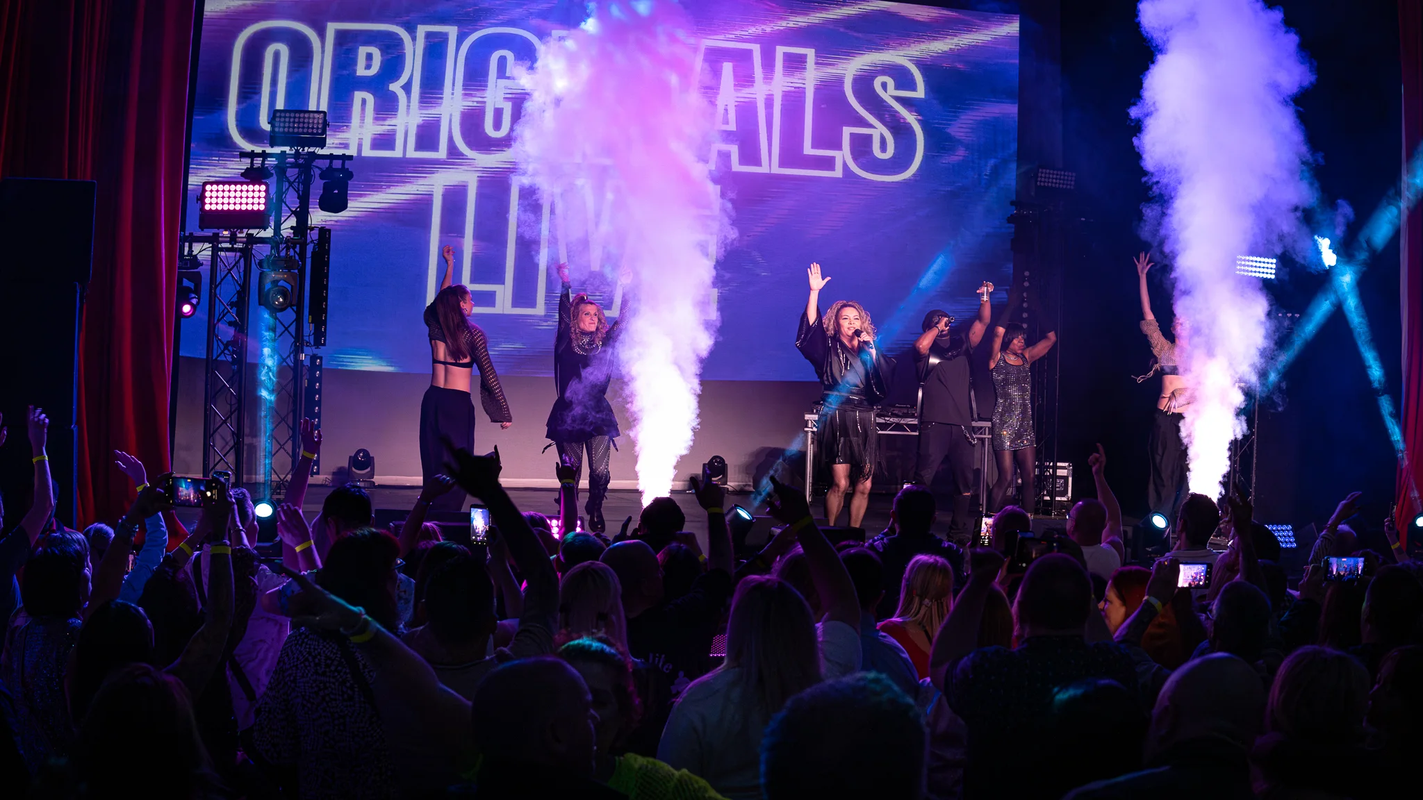 Live performers on stage with video, lighting and theatrical smoke in front of a packed audience